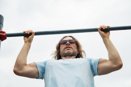 One fit young adult man or athlete in blue t-shirt training pull ups at sport public park. Sunny outdoor setting.の写真素材