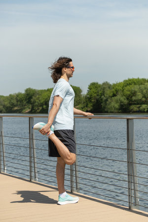 Fit brunette man wearing sports casual clothes doing stretching legs exercise on city modern outdoor...