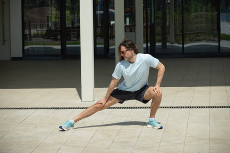Fit brunette man wearing sports casual clothes doing stretching and warm up exercise on urban...