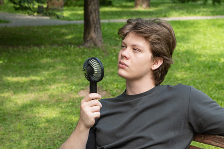 Handsome young caucasian man with hand electric fan suffering from heat at park. Heat Summer....