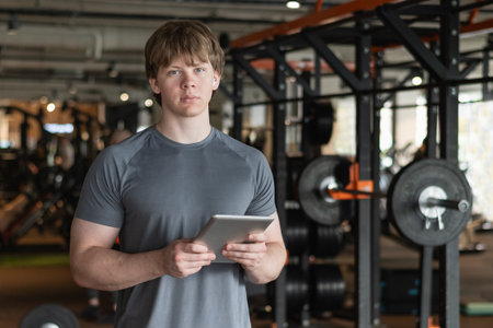 Serious personal trainer or bodybuilder standing wearing gray t-shirt holding tablet or clipboard in gym or fitness center. Coach making exercise plan in gymの写真素材