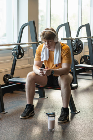 Young caucasian sporty man relax and sitting with mobile phone, protein shaker, listen music...