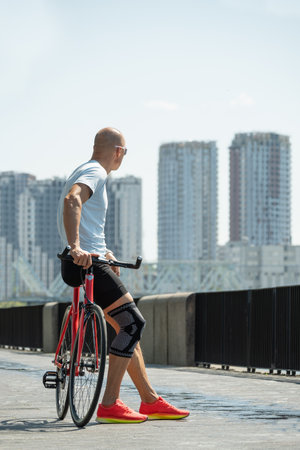 Cyclist pauses to enjoy city skyline on sunny day. Standing next to red bike, wearing athletic gear and sunglasses, he thoughtfully at modern buildings.の写真素材