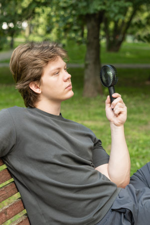 Handsome young caucasian man sitting relax on bench with hand electric fan suffering from heat at park. Heat Summer. Handheld fan. Beat heat.の写真素材