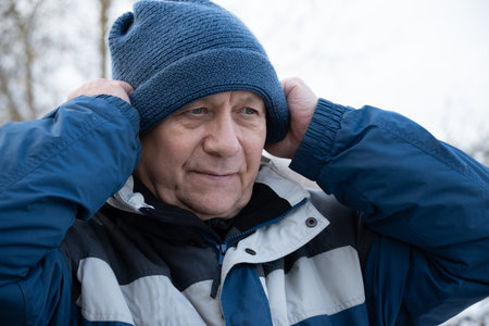 Senior man wearing blue winter knitted hat and jacket adjusts his hat outdoors. Man preparing for...