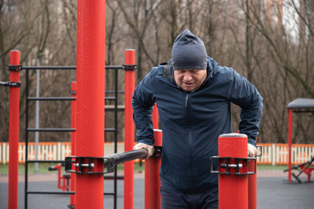 Focused man exercises on parallel bars in an urban outdoor gym after rain. Dressed warmly,...