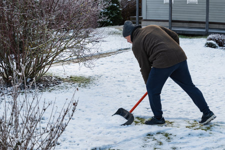 Senior man clearing snow in backyard with shovel, performing winter chores and maintaining clean...