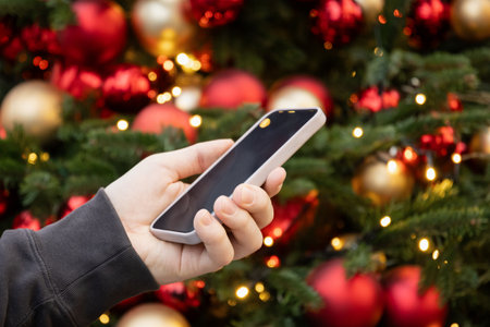Smartphone in mans hand against Christmas tree with decorative and light garland. Screen display for...