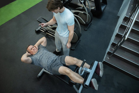 Adult man performs abdominal crunches under trainer guidance in a gym, strengthening core and...