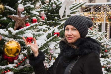 Woman holding a red Christmas ornament on a decorated fir tree at an outdoor holiday market.の写真素材