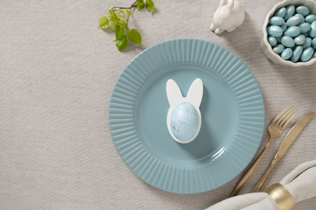 Pastel Easter table setting with blue plate, speckled egg in bunny holder and spring treats. Soft...