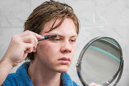 Young caucasian man using an electric trimmer to shape his eyebrows while looking into mirror in...
