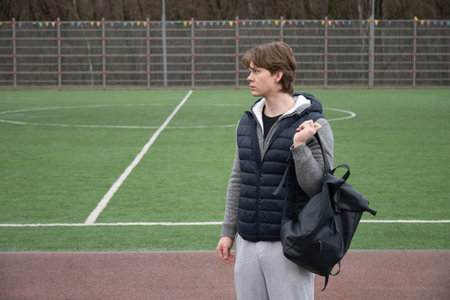 Walking across an outdoor sports field while carrying backpack, young adult caucasian man an...