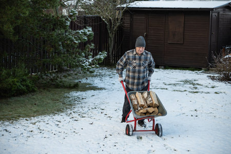 Man with firewood in wheelbarrow and snowy backyard. Winter firewood routine preparing and lighting...