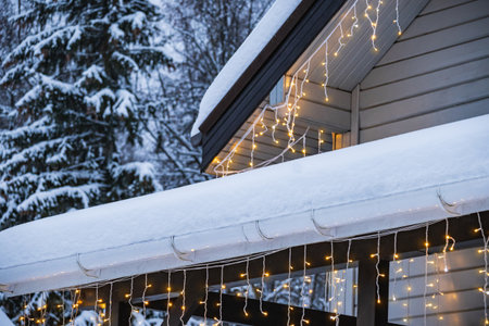 Close up of snow covered roof and porch with warm glowing Christmas string lights on winter house....