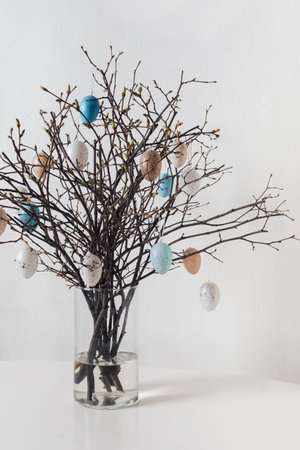 Decorative Easter branches with hanging pastel and speckled eggs in glass vase on white background....