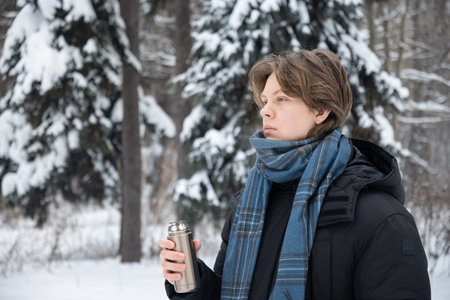 Young adult man holding metal thermos while standing in snowy forest. Winter outdoor lifestyle and...