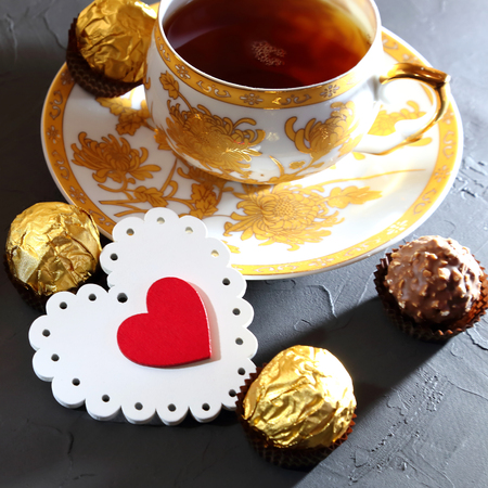Valentine's Day, a symbol of love. Still life - tea in a golden cup, delicate red and white heart and chocolate sweets on a black textured background.の写真素材