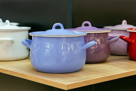 Many enameled steel metal multicolored new modern cooking pots stand on a shelf in the kitchen. Household goods, dishes, kitchen utensils. Shop window, sale of domestic products, interior detailsの写真素材