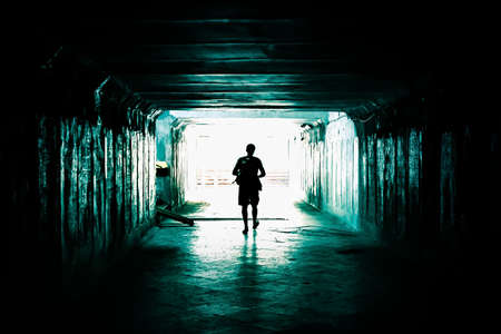 The silhouette of a man in the terrible mystical horror corridor of the basement with a paranormal ghostly light at the end of a dark tunnel with eerie shadows causing a feeling of fearの写真素材