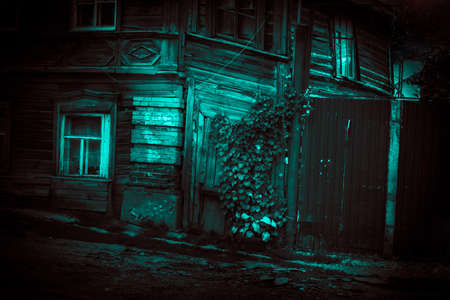 Old scary mystical abandoned wooden house with dark windows, fence and horror style wicket in dramatic paranormal ghostly blue green light of the moon with creepy strange shadows trees and ivyの写真素材