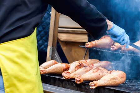 Close-up of a hands of man, dressed in a black shirt and in a yellow apron, cooking turkey legs on the hot grillの写真素材
