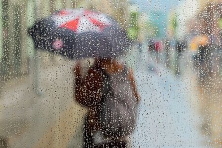 Abstract blurred silhouette of girl under umbrella, city street seen through raindrops on window...