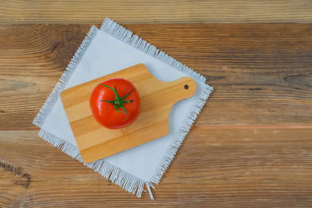 One fresh red ripe tomato with water drops on branch on wooden background with rustic wooden chopping board in center and white linen napkin. Top view, copy spaceの写真素材