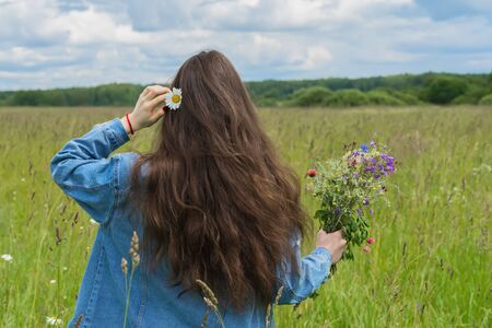 Unrecognizable young girl with daisie in her hair and beautiful summer bouquet of wild flowers in hand standing on background of green field. Seasons, environment, summer hobbies, recreation, lifestyleの写真素材