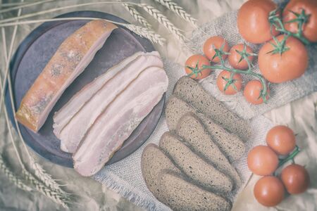 Rustic style of fresh organic food. Quick tasty snack. Slices of bacon, rye black bread, ripe red tomatoes on napkin from burlar. Top view. Vintage backdropの写真素材