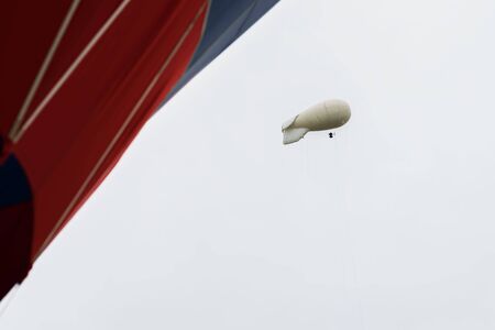 White coloured blimp, hot air airship in grey sky in rainy day nearby with vivid red Hot Air Balloon. Inflatable dirigible. Abstract modern background. With place for textの写真素材