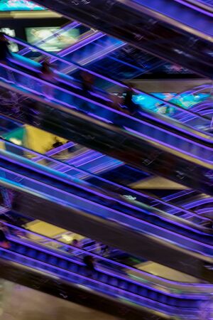 Abstract blurred image of modern shopping mall, unrecognizable silhouettes of people on escalators, purple led backlight, vertical background for design. Motion effectの写真素材