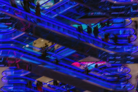 Abstract blurredn image of shopping mall, unrecognizable silhouettes of people on escalators with bright blue backlight, modern background for design. Motion effectの写真素材