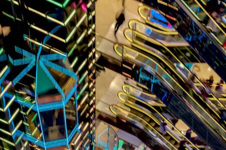 Abstract blurred image of modern shopping mall, unrecognizable silhouettes of people on escalators and glass elevator, bright vivid backlight. Motion effectの写真素材