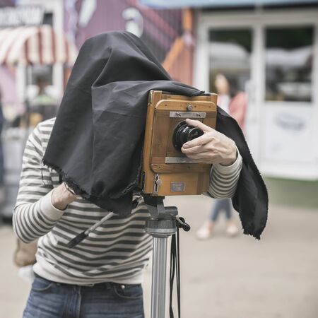Photographer with vintage wooden camera under dark cloth cape, photographing clients, artifact,...