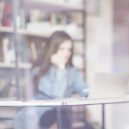 Abstract blurred bookcase in public library and young girl sitting at table, looking laptop, light background. Concept of learning, school, back to school, educationの写真素材