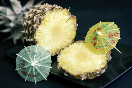 Slices of ripe pineapple on a black plate with decorations for cocktails, umbrella on dark...