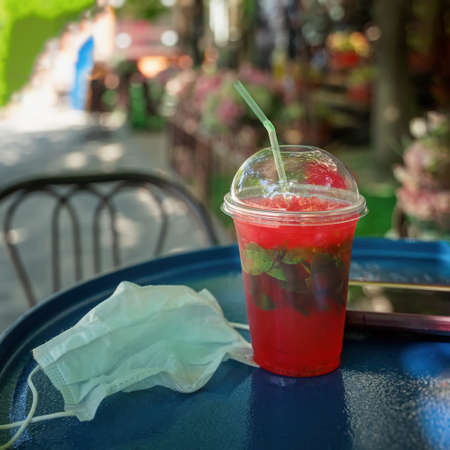Disposable plastic cup with refreshing drink and protective medical mask, Street cafe table,...