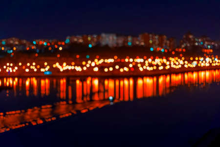 Abstract blurred bokeh of city, neon night lights, illumination, various bright colors in night with reflection in the lake. City life in evening, urban defocused backgroundの写真素材
