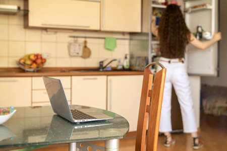 Remote work, home office. Laptop on the dining table, lunch break, unrecognizable girl takes food from the refrigeratordomestic, domestic lifestyleの写真素材