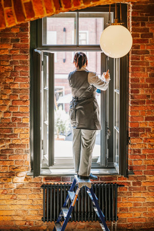 Woman worker washes window while standing with her back to us on stepladderの写真素材