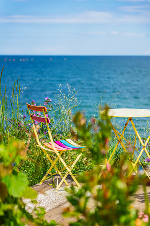 Bright yellow chair with multicolored stripes stands near a blue sea. Relaxing summer atmosphere with wildflowers and grass, scenic coastal settingの写真素材