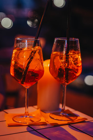 Two cocktail glasses with orange drink and straws on table in low light. Cocktail glow enhances nightlife, celebration, date vibe, cozy evening gathering outdoorsの写真素材