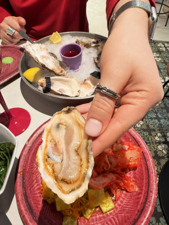 Womans hand holding oyster above vibrant plate with pickled vegetables during gourmet meal with seafood and wine. Concept of dining, seafood indulgence, flavor fusionの写真素材