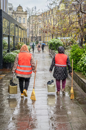 Women wearing safety vests cleaning wet city street during light snowfall. Bad weather, climate change, mud and puddles. Concept of outdoor public service, city maintenanceの写真素材