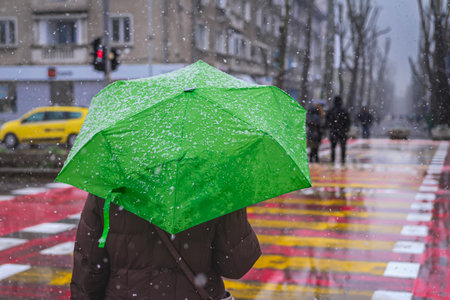 Person with green umbrella walking on city crosswalk during snowfall, urban winter scene. Bright green umbrella contrasts with snowy cityscape. Concept of winter, city life, seasonsの写真素材