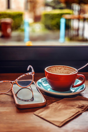 Red coffee cup with spoon on saucer near smartphone, eyeglasses on table by window. Warm indoor...