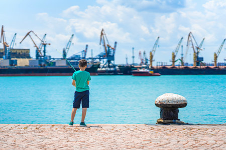 Boy fishing on sunny pier with industrial port in background, standing near turquoise water. Concept of fishing, childhood moments, peaceful outdoor leisureの写真素材