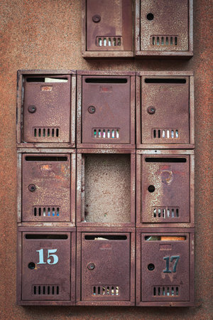 Old rusted metal mailboxes with peeling numbers and missing compartments attached to textured urban wall. Concept of urban nostalgia and vintageの写真素材