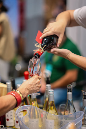 Hand pouring red wine into glass during tasting event, surrounded by bottles and vibrant atmosphere...